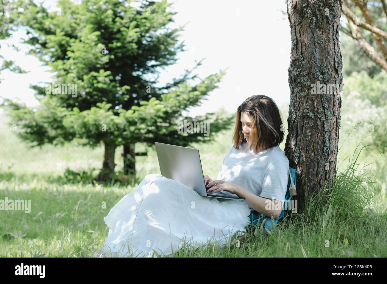 Nettes Teenager-Mädchen, das in der Nähe von Baum im Sommerpark mit offenem Laptop in den Händen sitzt und studiert. Konzept des Fernlernens Stockfoto