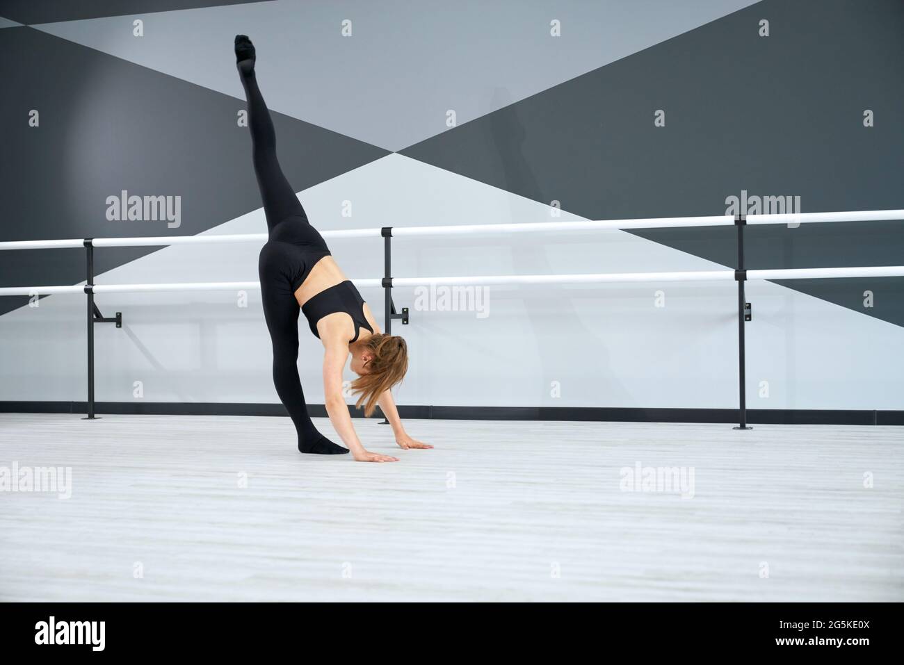 Seitenansicht von stark fit Mädchen in engen schwarzen Sport-Outfit Stretching und üben Split in der Luft. Flexible Tänzerin, die im Tanzsaal mit den Handläufen des Balletts den Boden berührt. Sportkonzept. Stockfoto