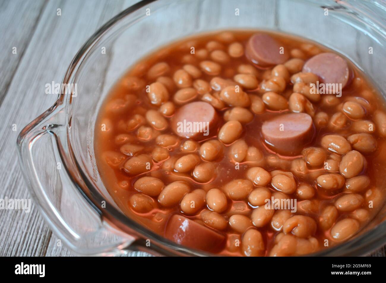 Franks und Bohnen oder Beanie-Weenies in einer klaren Glasschale auf einer Holzoberfläche Stockfoto
