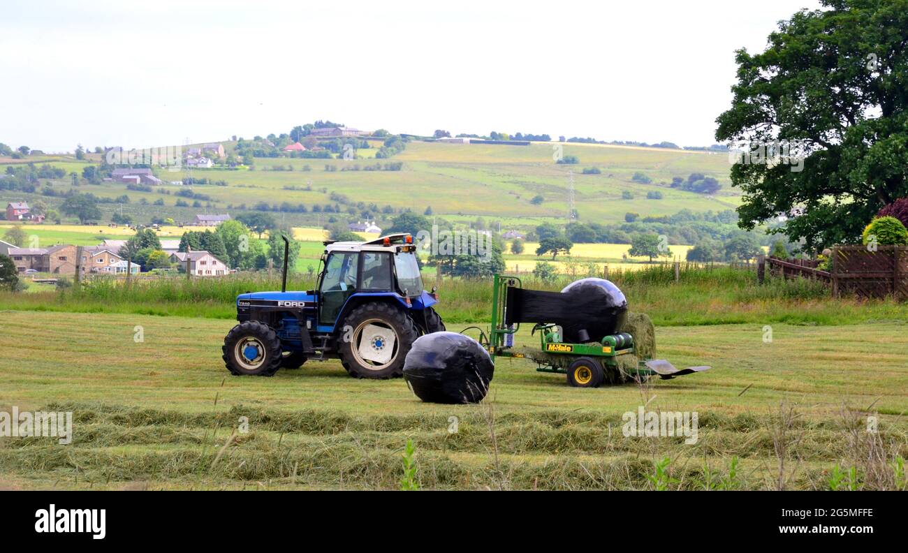 Traktor mit Rundballengrassilage-Verpackungsmaschine aus Kunststoff bei der Arbeit auf dem Feld im Großraum Manchester, Großbritannien Stockfoto