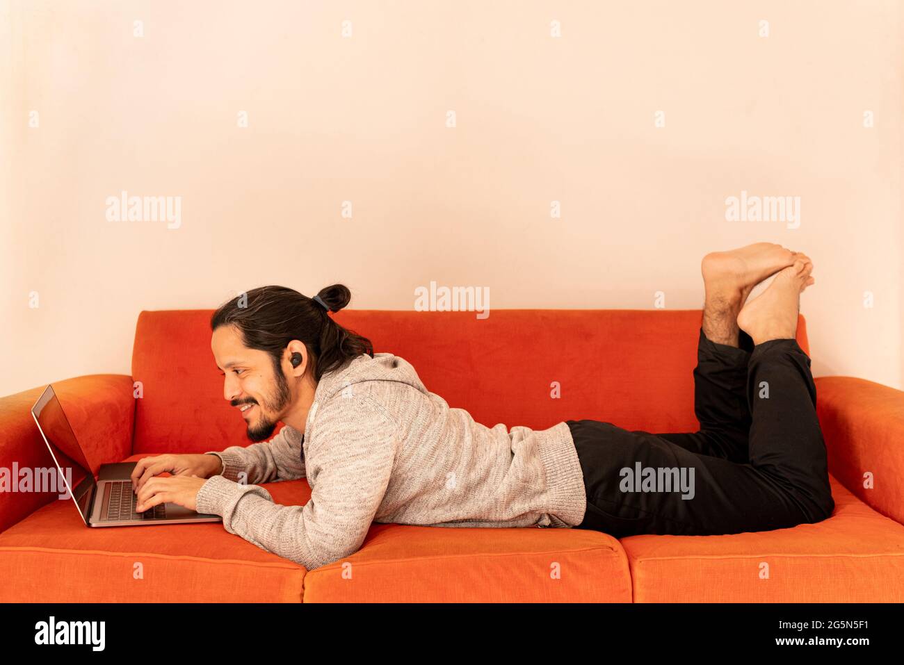 Junger Mann, der online mit dem Computer-Laptop unterwegs ist, der auf einem orangefarbenen Sofa zu Hause im Wohnzimmer liegt. Stockfoto