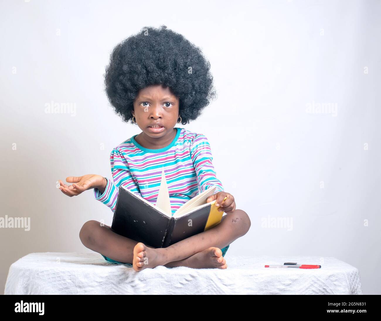 Afrikanisches Mädchen mit afro-Frisur zeigt ihre Liebe zur Bildung, während sie sitzt und einen Stift hält und auch ein Buch auf dem Schoß hat Stockfoto