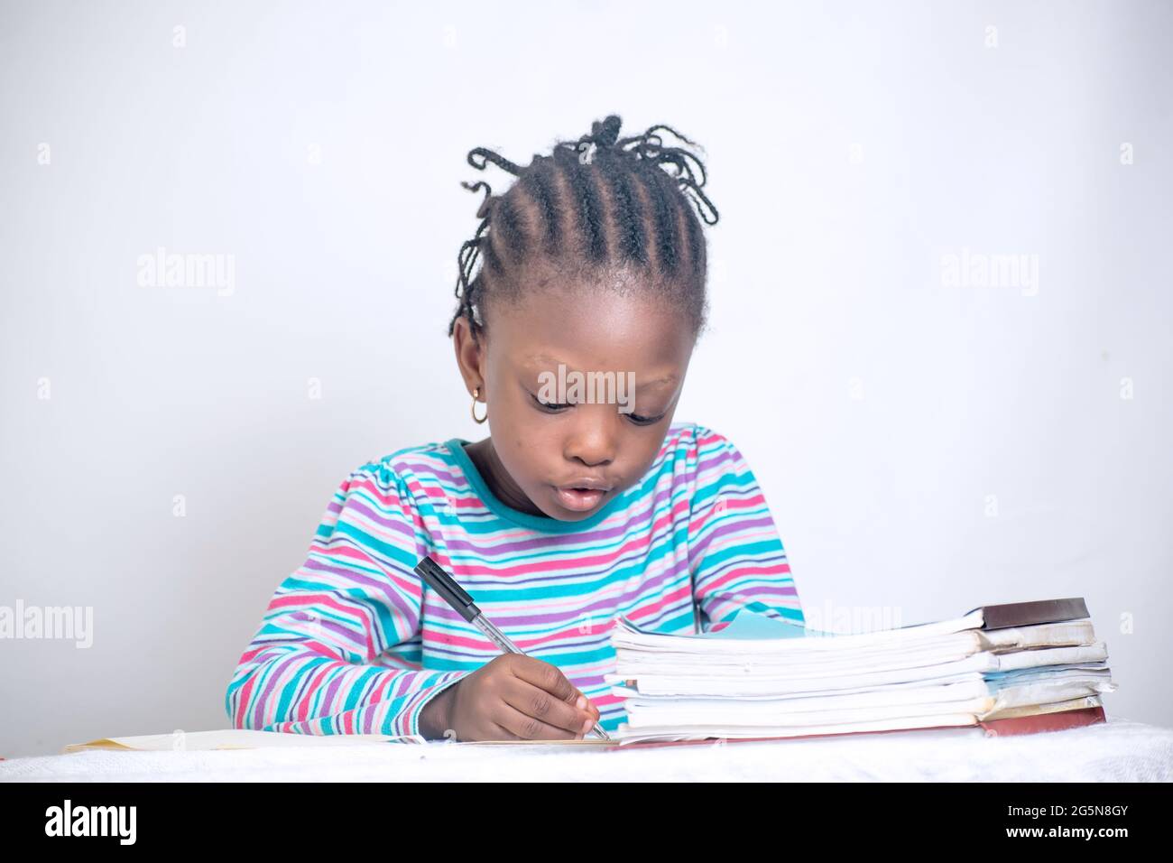 Nahaufnahme eines Bildungstisches eines afrikanischen Mädchens mit gewebter Frisur, das auf einem Lerntisch schreibt, während es Bücher und einen Blumenkorb von ihr hat Stockfoto