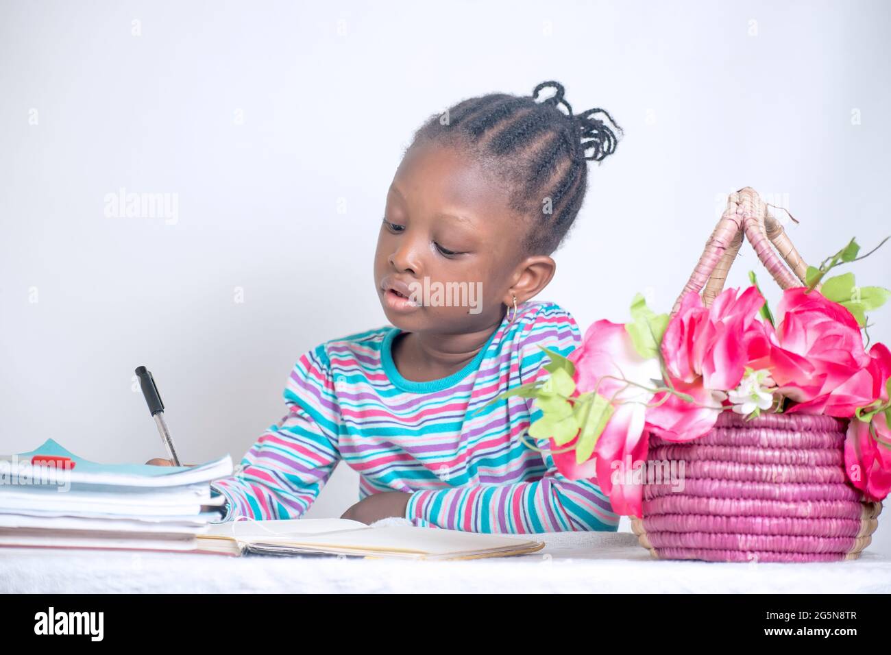 Nahaufnahme eines Bildungstisches eines afrikanischen Mädchens mit gewebter Frisur, das auf einem Lerntisch schreibt, während es Bücher und einen Blumenkorb von ihr hat Stockfoto
