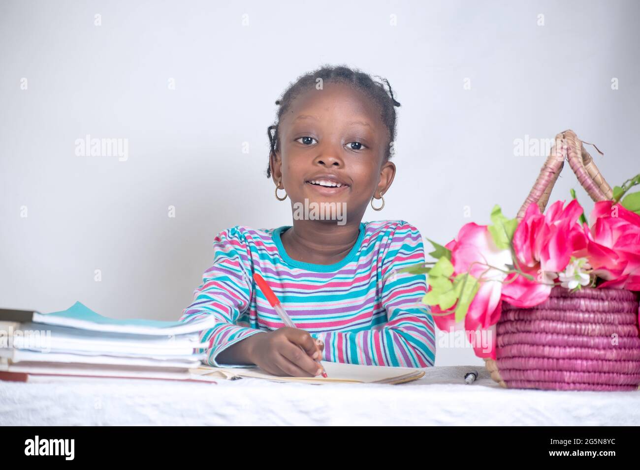 Nahaufnahme eines Bildungstisches eines afrikanischen Mädchens mit gewebter Frisur, das auf einem Lerntisch schreibt, während es Bücher und einen Blumenkorb von ihr hat Stockfoto