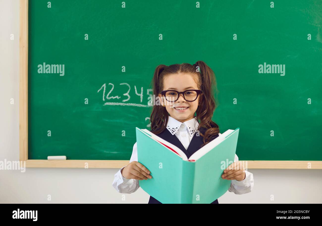 Lustige kleine Schülerin in Brille stehend mit Buch in einer Lektion in der Klasse in der Schule. Stockfoto