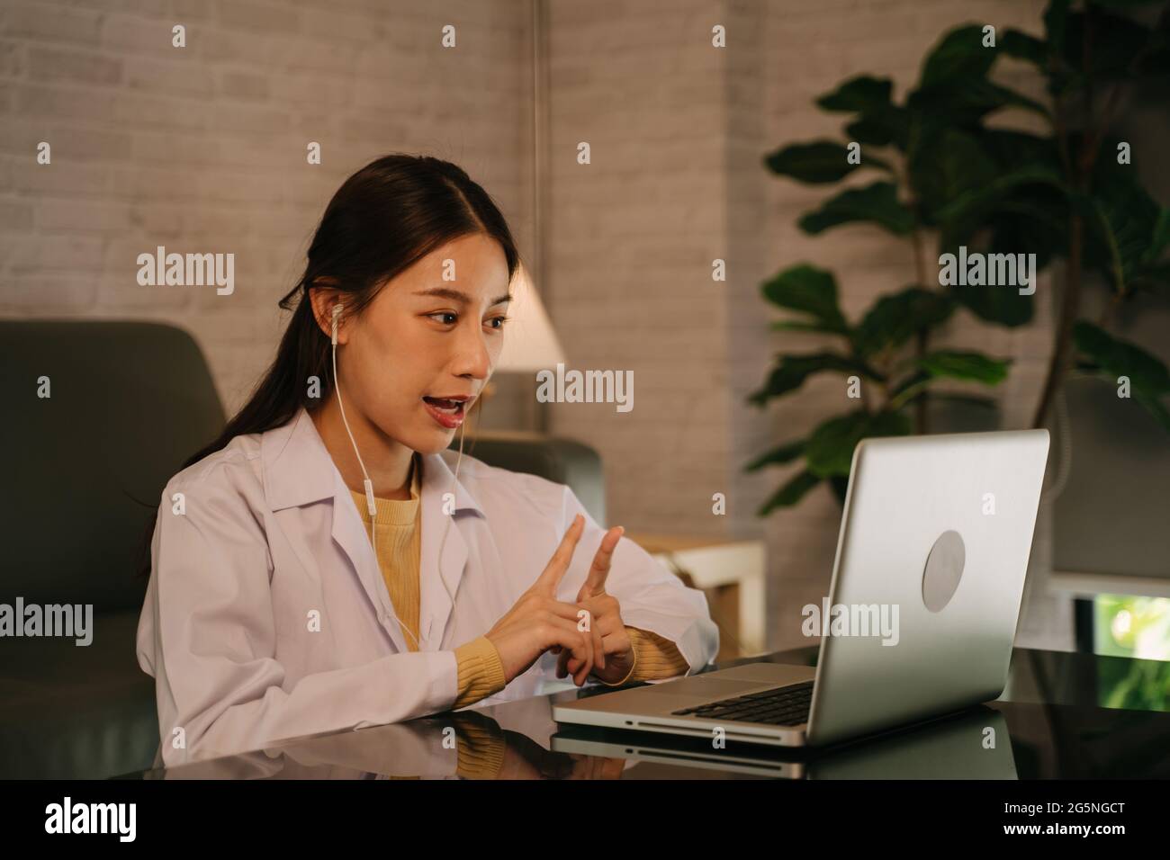 Erfolgreiche junge asiatische Ärztin mit Laborkittel und Ohrhörern, die in der späten Nacht Videokonferenzgespräche zur Konsultation mit Patienten von zu Hause aus führt Stockfoto