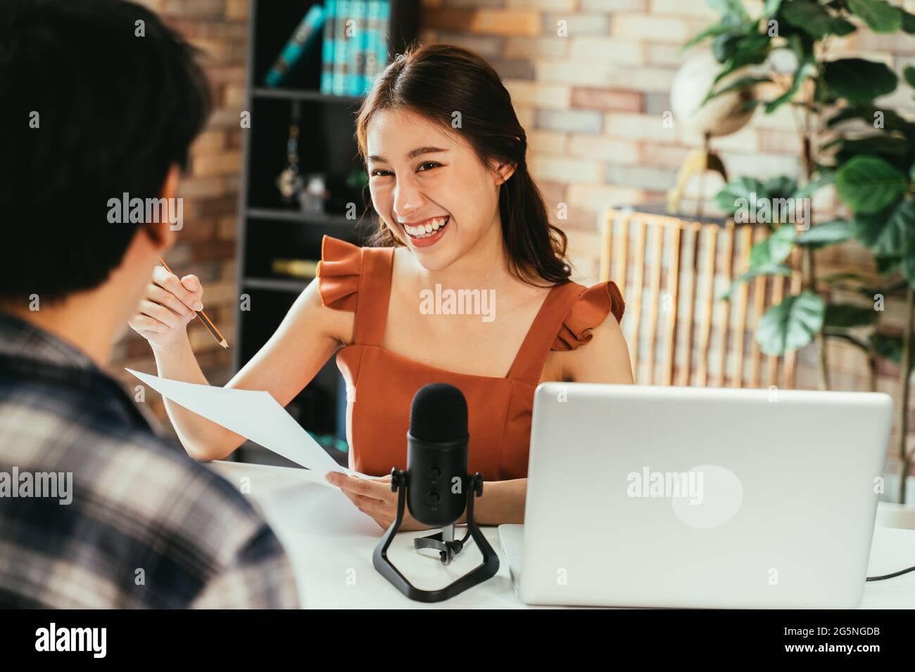 Fröhliche junge asiatische weibliche Podcasterin hält einen Fragebogen, während sie männliche Gäste am Arbeitsplatz mit Mikrofon und Laptop interviewt Stockfoto