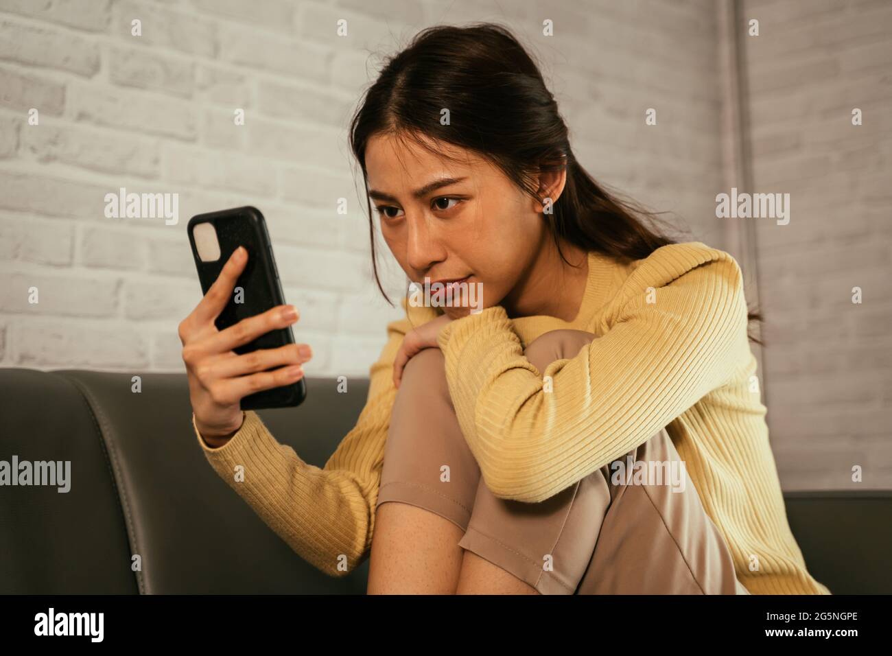 Junge und müde asiatische Frau sitzt ängstlich und nervös auf der Couch zu Hause Blick auf Handy, während sie auf einen Anruf oder Nachrichten warten Stockfoto