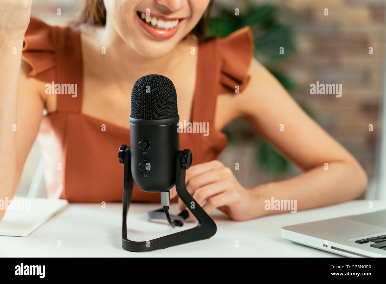 Nahaufnahme einer jungen, schönen Frau, die lächelnd auf dem Schreibtisch sitzt und dabei einen Podcast mit Laptop und Mikrofon aufzeichnet, während sie ein Interview führt Stockfoto