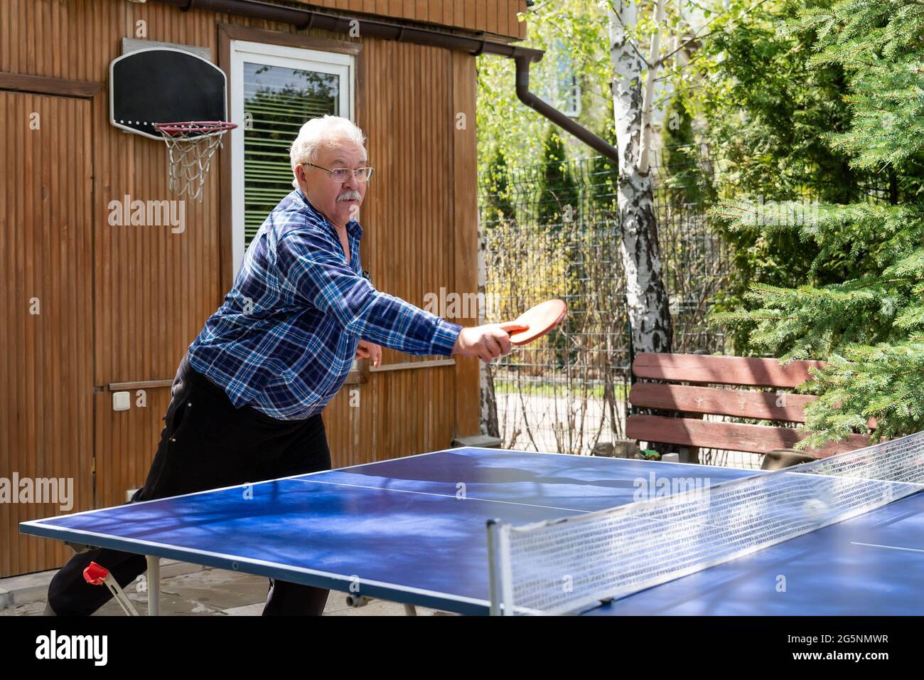 Ältere ältere graue kaukasische alte männliche Person, die starke Kugel geschossen spielt Tischtennis im Garten des Hauses im Freien am sonnigen Tag. Ältere Menschen Stockfoto