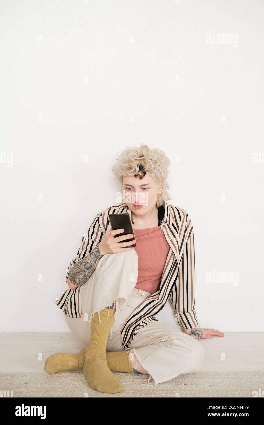 Junge Frau in stilvoller Kleidung, die auf dem Boden im Zimmer sitzt und ihr Mobiltelefon benutzt Stockfoto