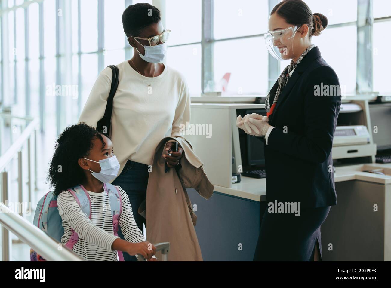 Mutter und Tochter mit Gesichtsmasken, die am Check-in-Schalter am Flughafen vorbeikommen und das Flughafenpersonal einen Gesichtsschutz trägt. Zweiköpfige Familie, die während Pandemi unterwegs war Stockfoto