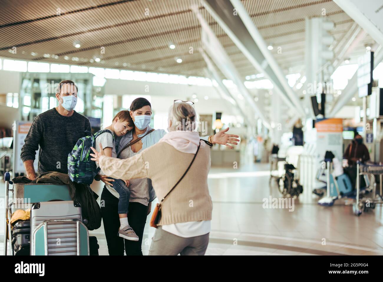 Eine ältere Frau begrüßt ihre Tochter und ihre Familie mit offenen Armen am Ankunftstor des Flughafens. Dreiköpfige Reisende Familie von Großmutter am Flughafen in Pan begrüßt Stockfoto