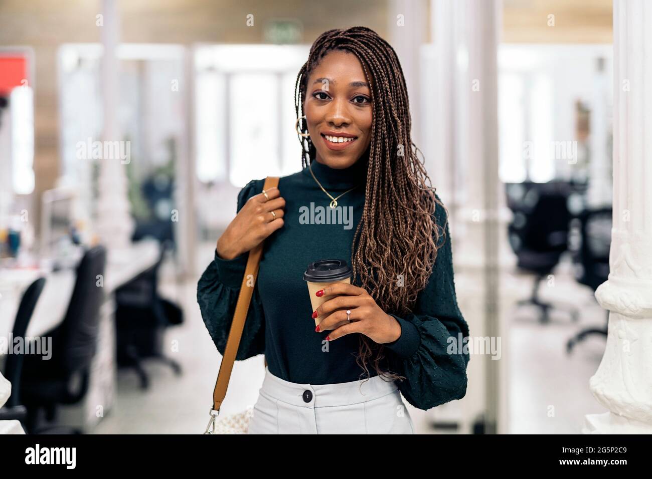 Schwarze Frau mit coolen Zöpfen und in formeller Kleidung lächelnd und mit Blick auf die Kamera. Sie arbeitet im Büro. Stockfoto