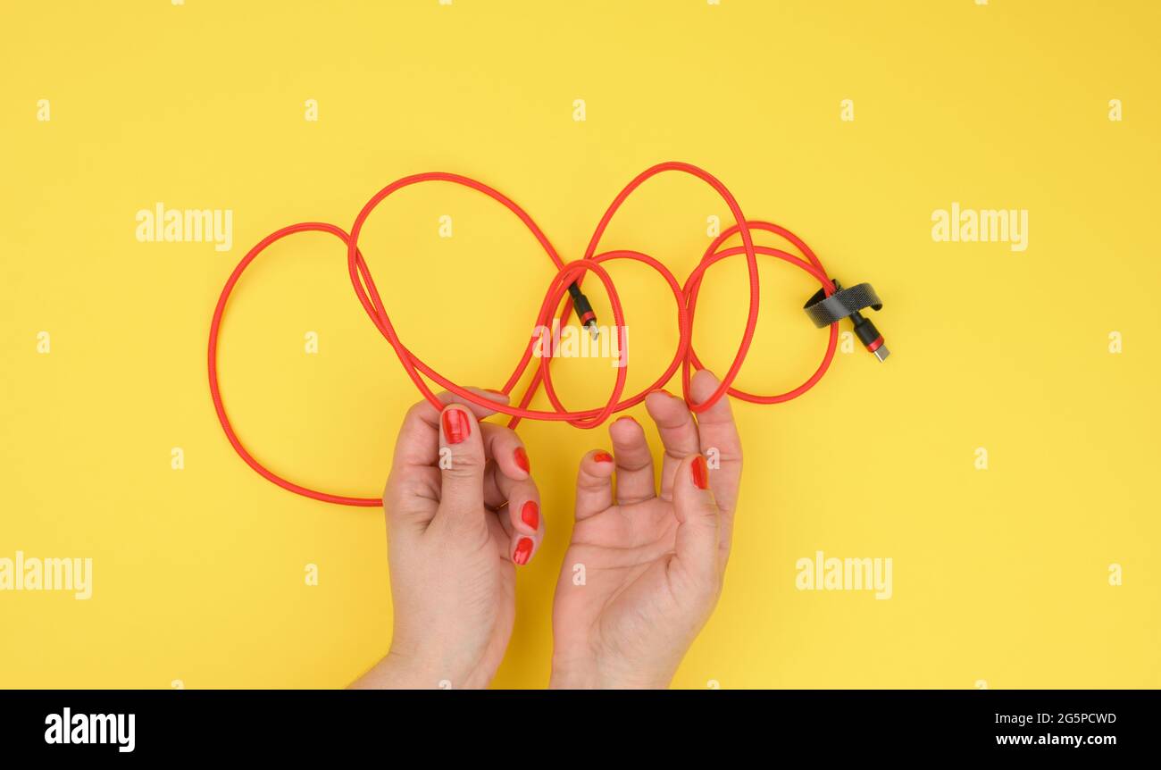 Weibliche Hand hält verdrilltes Kabel zum Laden von mobilen Geräten in einer roten Textilverpackung auf einem gelben Hintergrund, Nahaufnahme Stockfoto