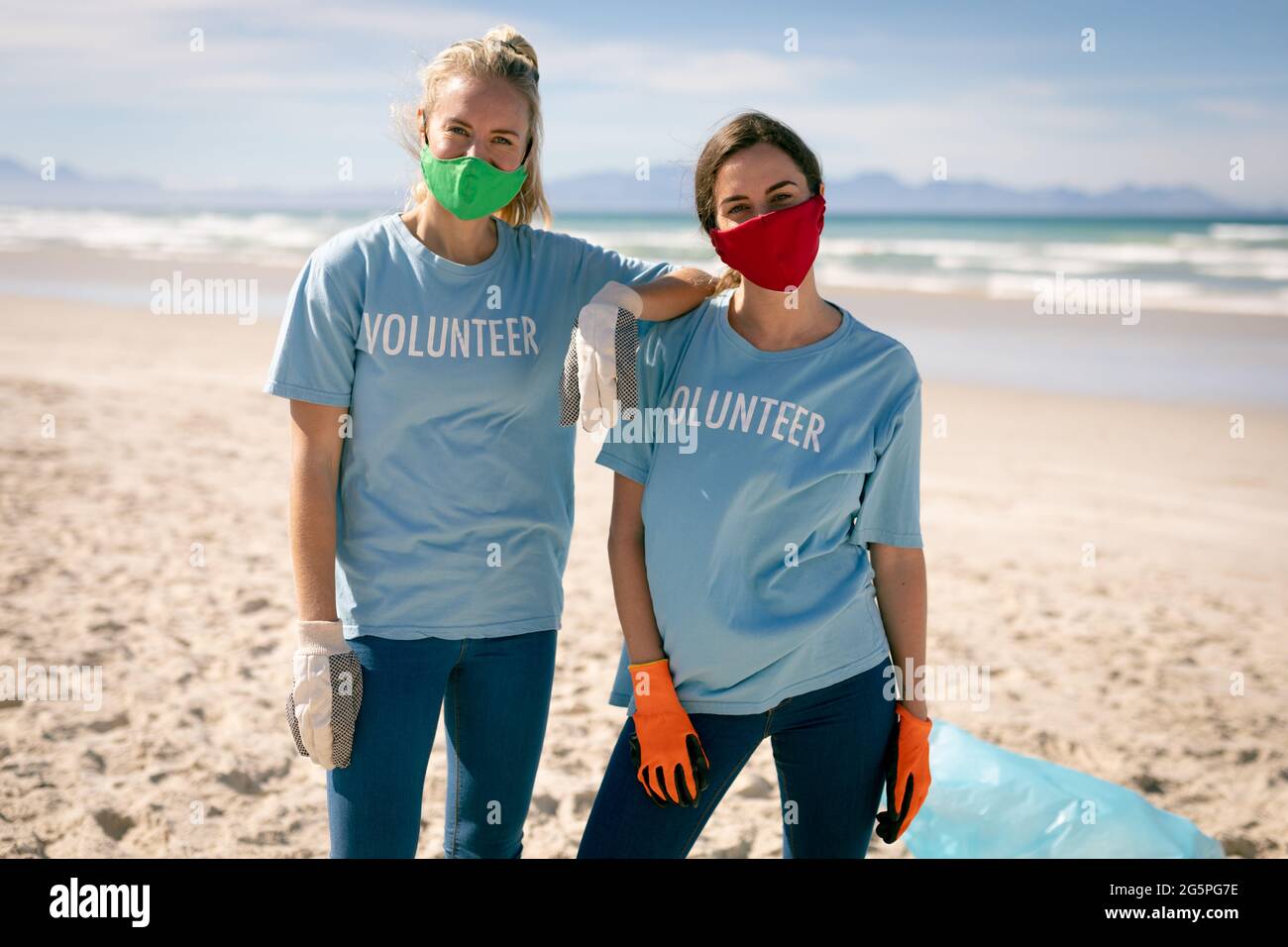 Zwei verschiedene Frauen tragen freiwillige T-Shirts und Gesichtsmasken, die Müll vom Strand aufsammeln Stockfoto