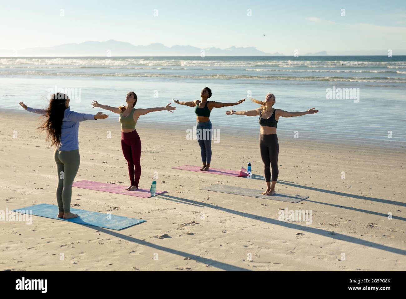 Eine vielfältige Gruppe von Frauen, die Yoga praktizieren und mit ausgestreckten Armen am Strand stehen Stockfoto