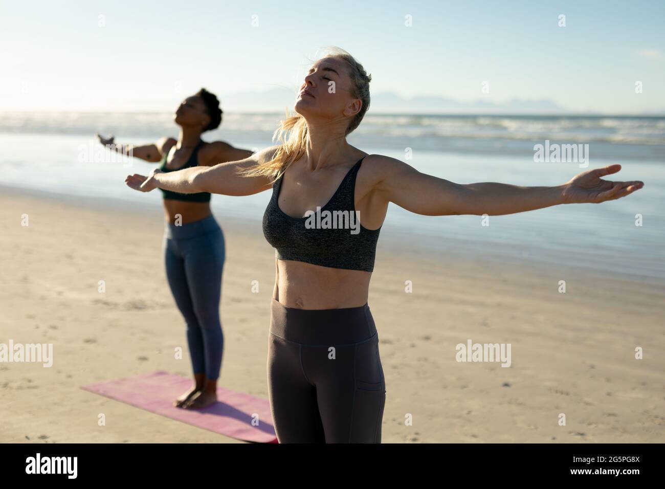 Zwei verschiedene Frauen, die Yoga praktizieren, stehen mit ausgestreckten Armen und meditieren am Strand Stockfoto