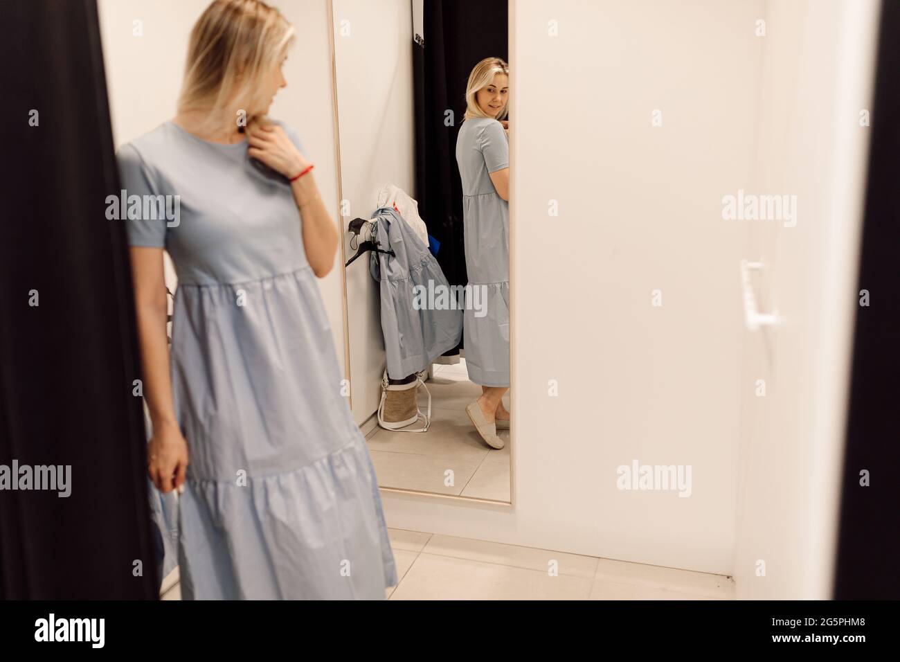 Moderne junge Frau hat die Möglichkeit zu beurteilen, wie sie Kleid in Umkleideraum des Bekleidungshauses trägt. Organisation des Verkaufsprozesses in Supermarke Stockfoto