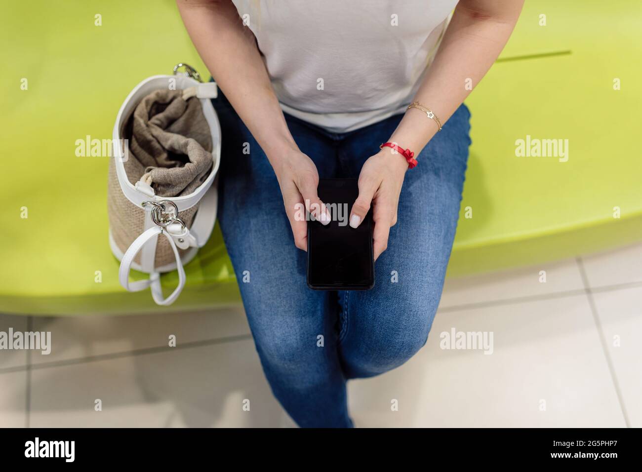 Die junge Frau sitzt auf dem hellgrünen Sofa im Einkaufszentrum und schaut auf ihr Telefon. Daneben steht eine Einkaufstasche. Moderne Technologien. Shopp Stockfoto