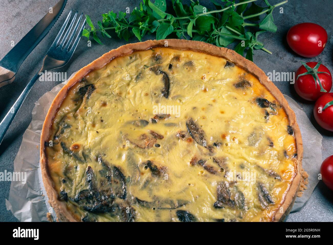 Nahaufnahme einer frisch gebackenen gelben Quiche mit Pilzen noch auf einem Backpapier, sitzend auf einer dunklen Küchentischoberfläche mit Besteckset, t Stockfoto