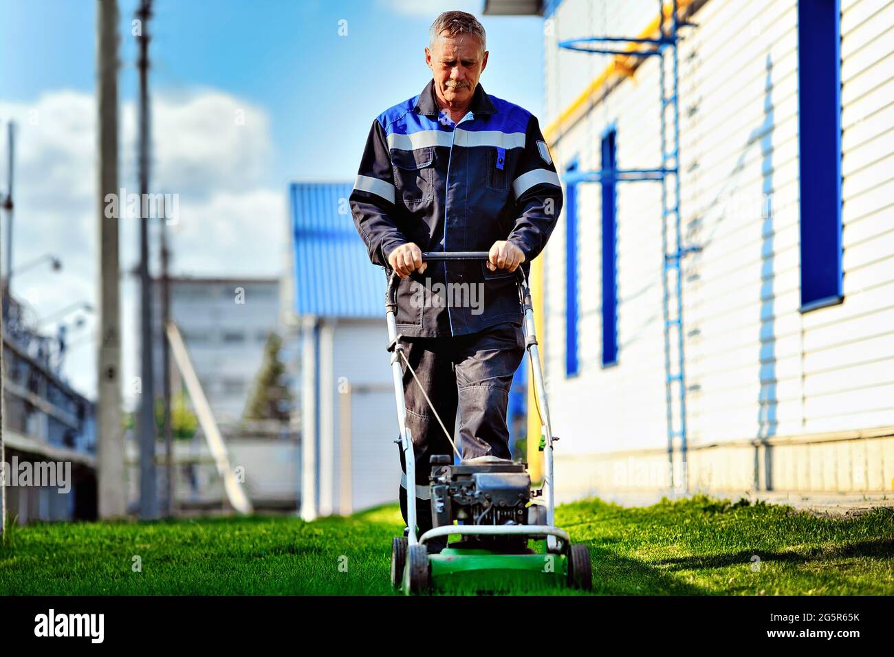 Ein Rentner in Arbeitskleidung arbeitet an einem sonnigen Tag mit einem Rasenmäher auf dem Gebiet der Produktionsbasis. Körperliche Arbeit und Teilzeitarbeit. Vorderansicht. Stockfoto