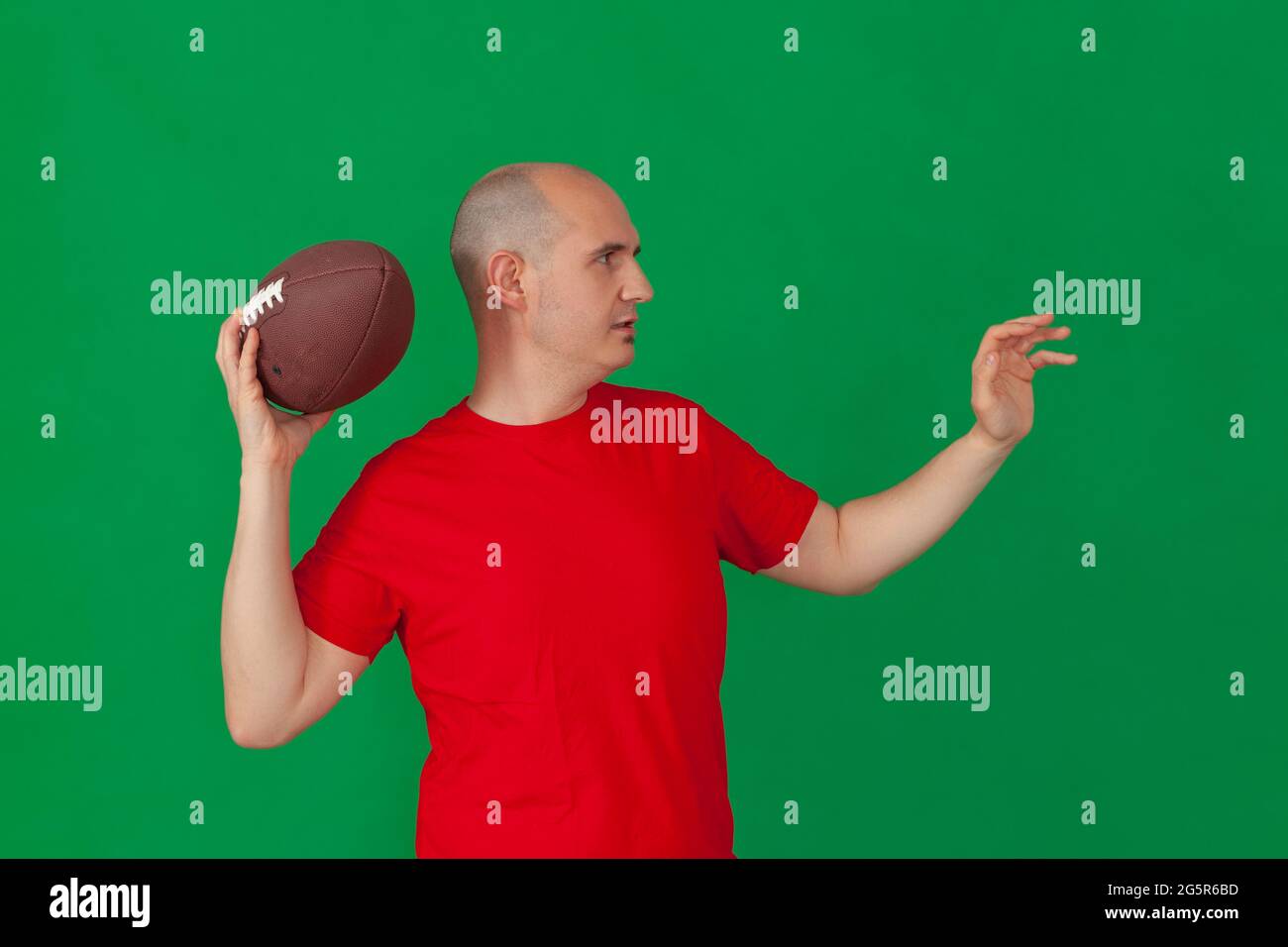 Ein kahlköpfiger Kaukasusmann, der in einem roten T-Shirt gekleidet ist und einen amerikanischen Fußballball hält, der so positioniert ist, als würde er einen Vorwärtspass werfen. Der Hintergrund ist grün. Stockfoto