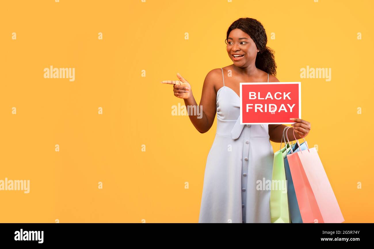 Charmante schwarze Frau mit Einkaufstaschen, die schwarzes FREITAGSSCHILD halten und auf leeren, orangefarbenen Hintergrund zeigen Stockfoto