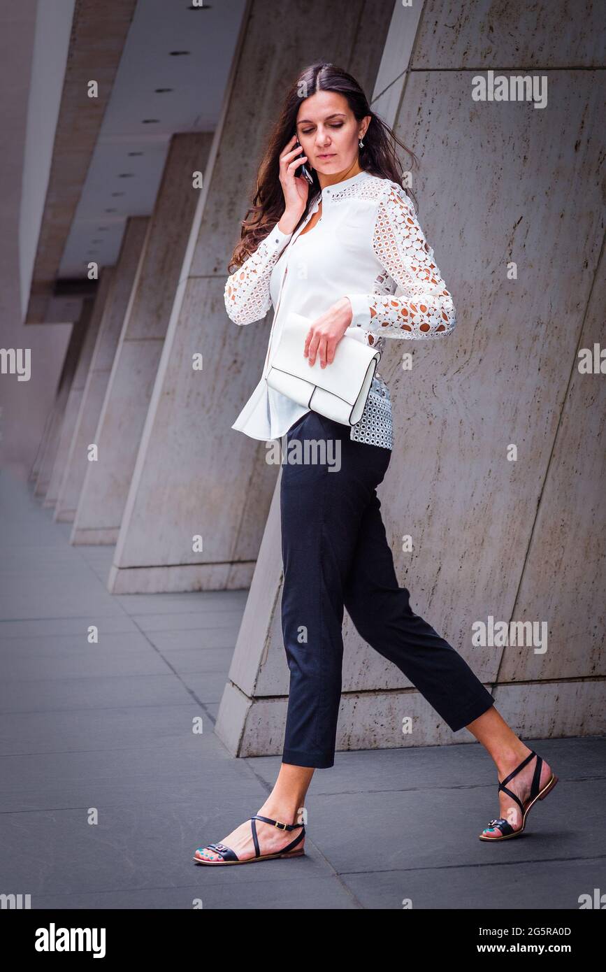 Eine junge osteuropäische Geschäftsfrau, die ein weißes, mit Spitze ausgeschnittenes Langarmhemd, schwarze Hosen, Sandalen und eine weiße Ledertasche trägt, ist Talki Stockfoto