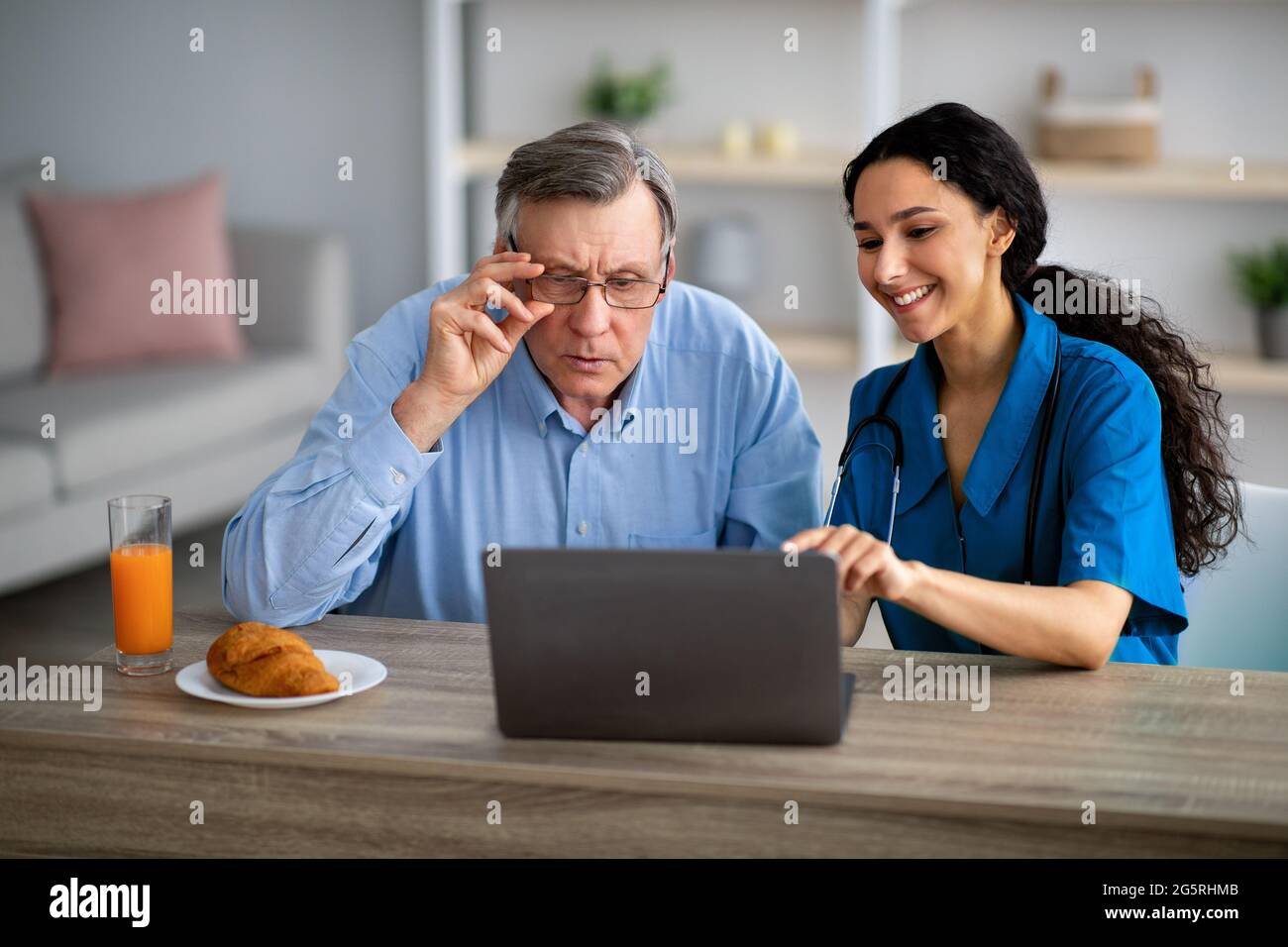 Die tausendjährige Pflegekraft zeigt älteren behinderten Menschen im Rollstuhl, wie sie den Laptop im Altersheim benutzen Stockfoto