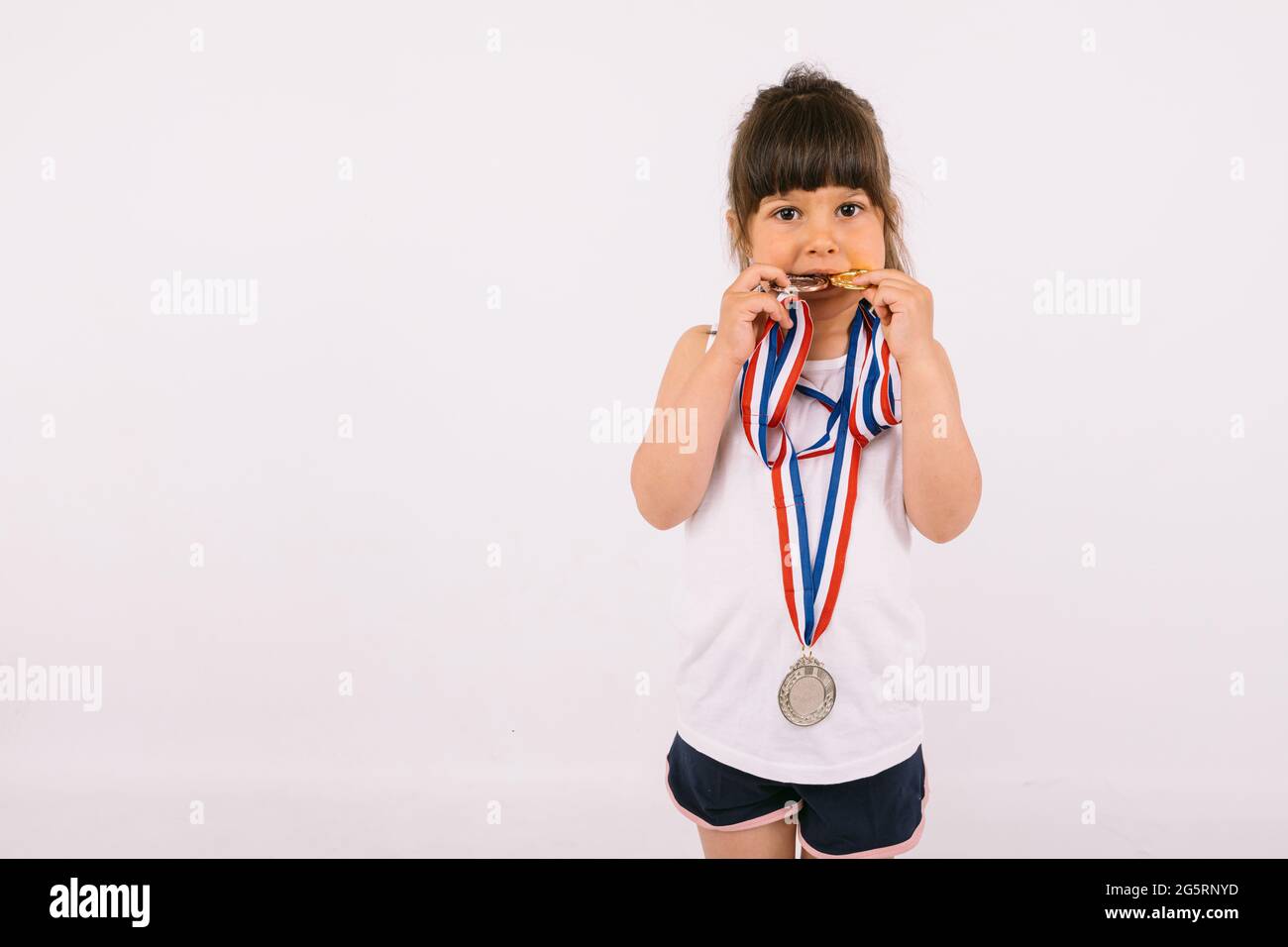 Kleines braunhaariges Mädchen mit Sportmeistermedaillen, das sie beißt. Sport- und Siegeskonzept Stockfoto