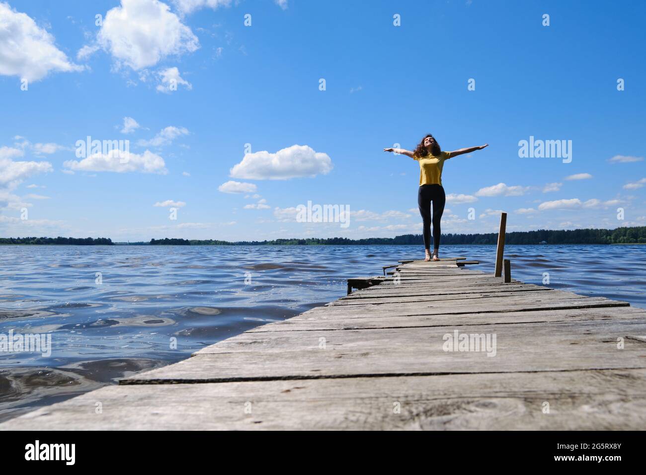 Mädchen, das am Pier in der Nähe des Flusses steht, ihre Hände nach oben hebt, genießt das sonnige Wetter. Stockfoto