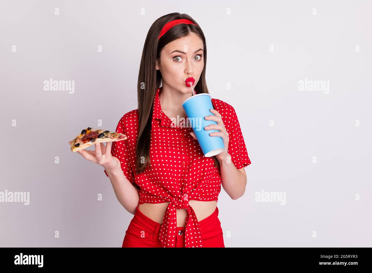 Foto von niedlichen schönen Mädchen halten Pizza Scheibe trinken Tasse Soda Stroh tragen rot gepunktete Bluse isoliert grauen Hintergrund Stockfoto