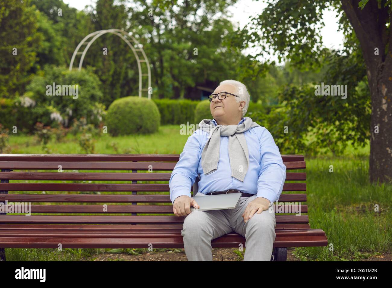 Ein glücklicher älterer Mann mit einem Laptop sitzt auf einer Bank in einem grünen Sommerpark Stockfoto