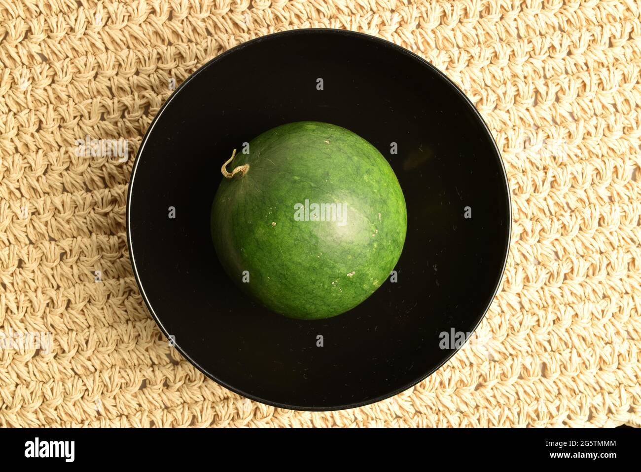 Eine dunkelgrüne Wassermelone auf einem schwarzen Teller, Nahaufnahme, auf einer Strohmatte, Draufsicht. Stockfoto