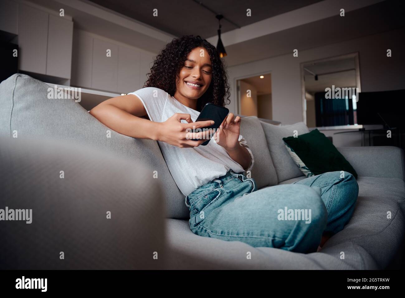 Mixed-Race-Frau lächelt beim Entspannen auf der Couch beim Tippen auf dem Mobiltelefon Hochwertige 4k-Aufnahmen Stockfoto