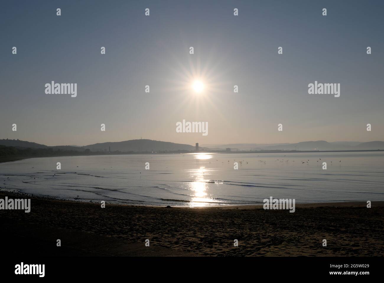 An einem klaren, blauen Tag geht die Sonne hinter Swansea City von der anderen Seite der Bucht auf Stockfoto