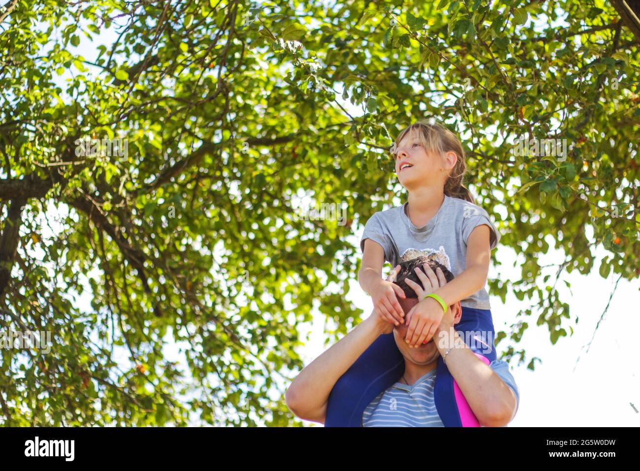 Defocused Bruder Reiten Schwester auf dem Rücken. Portrait von glücklichen Mädchen auf Mann Schultern, Huckepack. Mädchen fliegen. Familie spielt draußen. Grüner Baum verschwimmt Stockfoto