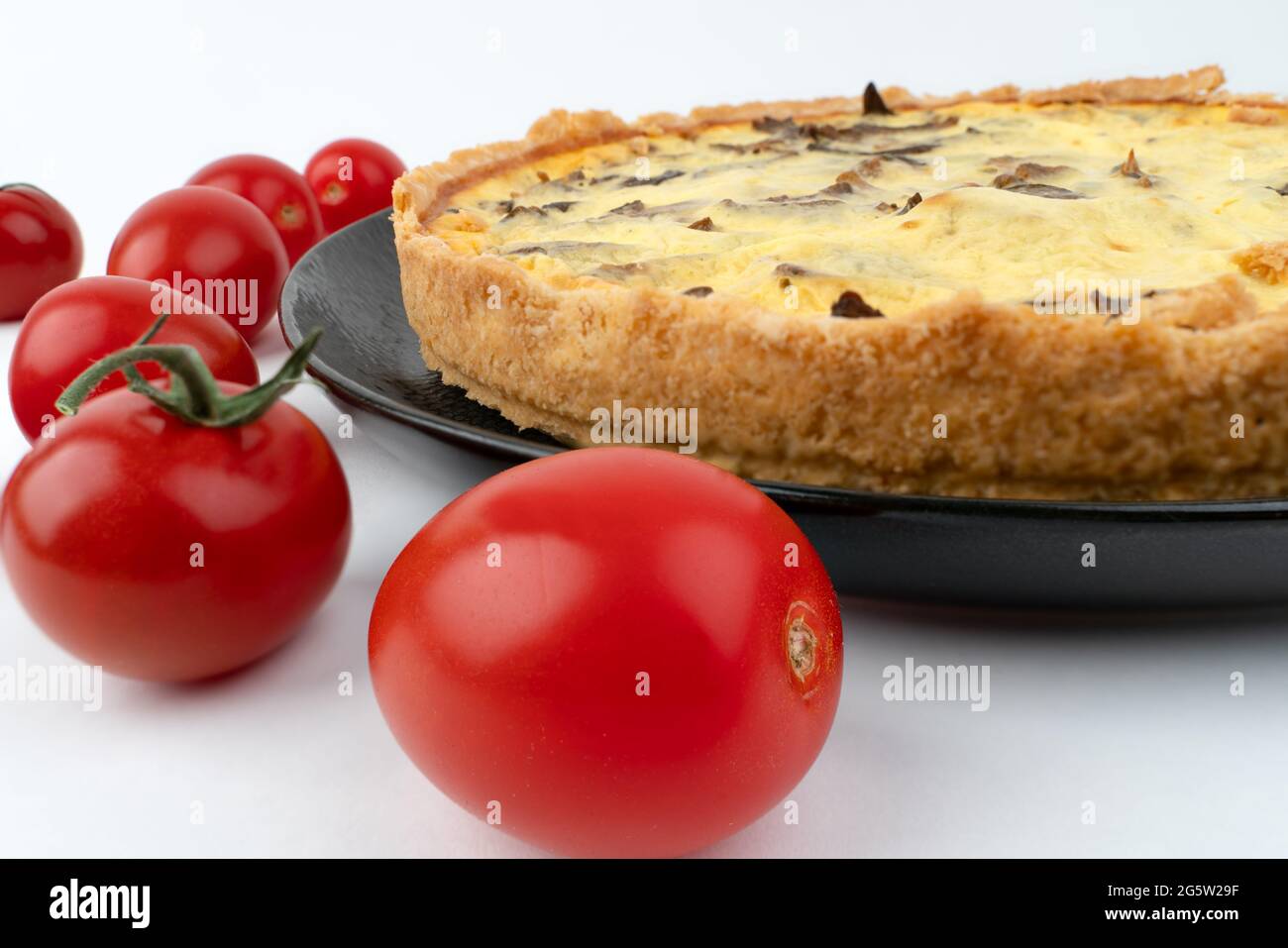 Nahaufnahme große Studioaufnahme von frisch gebackenen gelben französischen salzigen Kuchen oder Quiche, mit Pilzen, roten Kirschtomaten, auf einem schwarzen Design-Teller, isoliert Stockfoto
