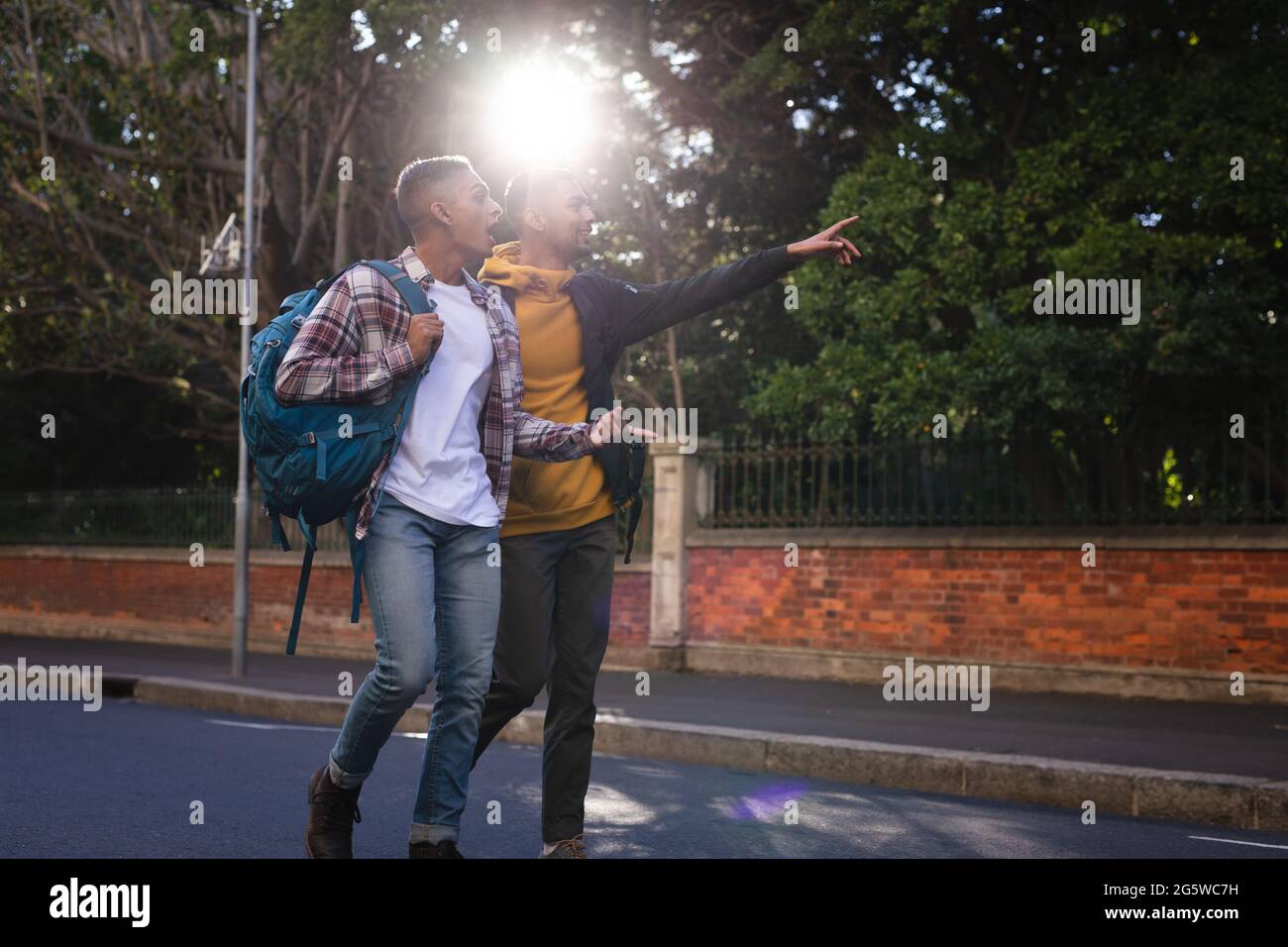 Zwei glückliche männliche Freunde mit gemischtem Rennen, die Rucksäcke tragen und in der Stadtstraße spazieren gehen, einer zeigt Stockfoto