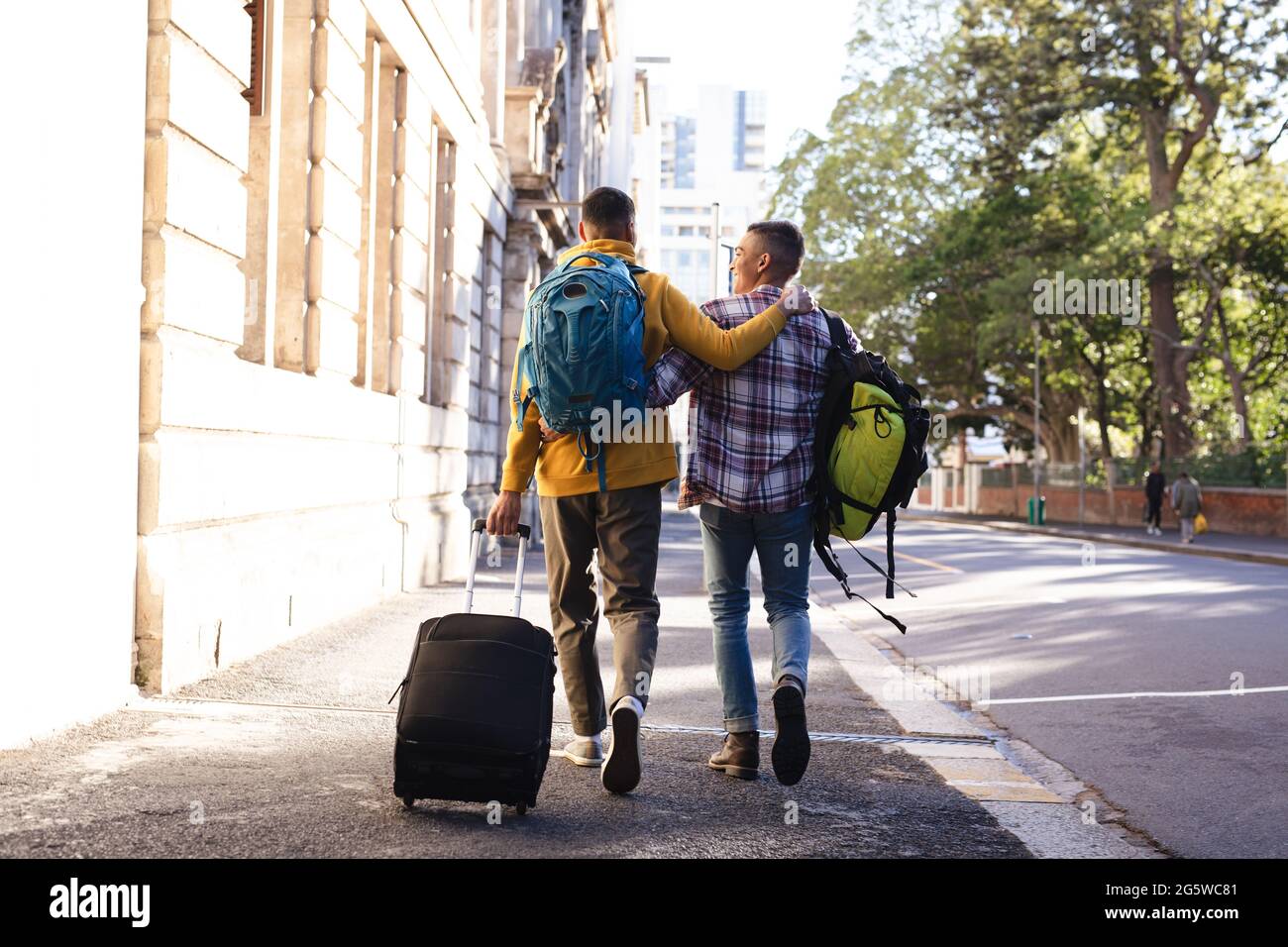 Rückansicht von zwei männlichen Mischrennfreunden in der Stadtstraße, die umarmt entlang gehen und Gepäck tragen Stockfoto