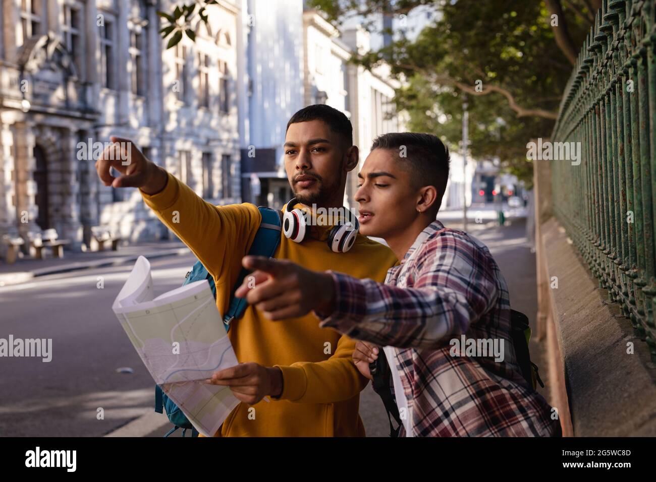Zwei männliche Freunde mit gemischter Rasse, die in der Stadtstraße stehen, auf die Karte schauen, sprechen und in die Richtung zeigen Stockfoto