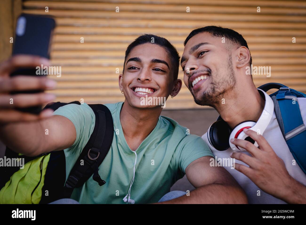 Zwei fröhliche, gemischte männliche Freunde mit Rucksäcken, die in der Stadtstraße sitzen und Selfie machen und lächeln Stockfoto
