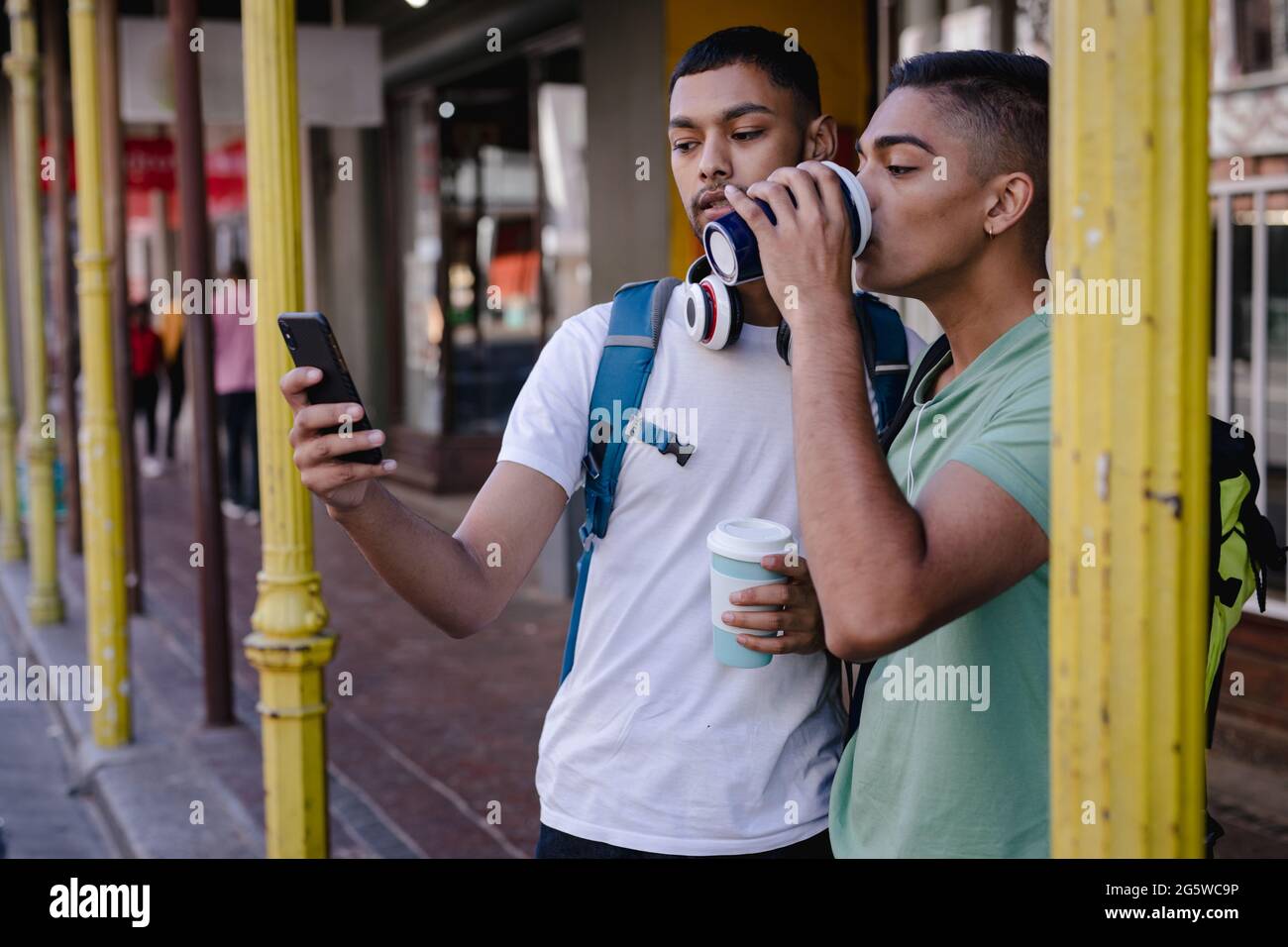 Zwei männliche Mischrennfreunde mit Rucksäcken, die in der Stadtstraße stehen und Kaffee trinken, mit dem Smartphone Stockfoto
