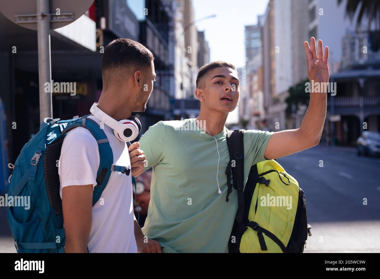 Zwei männliche Mischrennfreunde mit Rucksäcken, die in der sonnigen Stadtstraße stehen, ein Taxi Stockfoto
