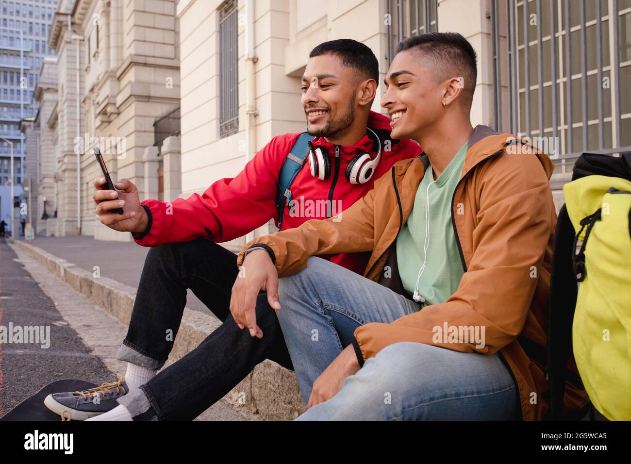 Zwei glückliche gemischte Rennen männliche Freunde mit Rucksäcken sitzen in der Stadt Straße unter Selfie Stockfoto