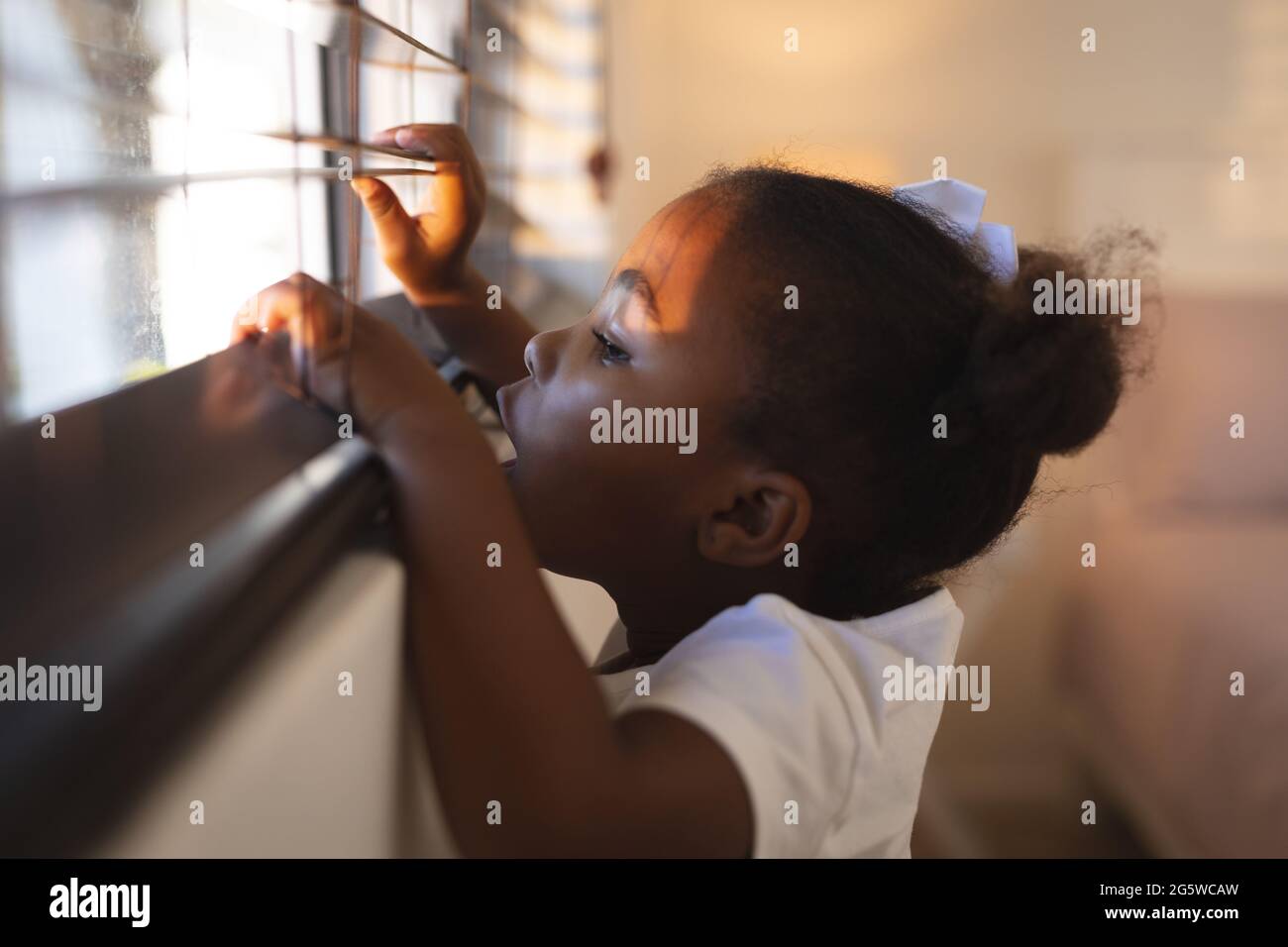 Neugieriges afroamerikanisches Mädchen, das an einem sonnigen Tag durch die Jalousien gucken und stehen Stockfoto