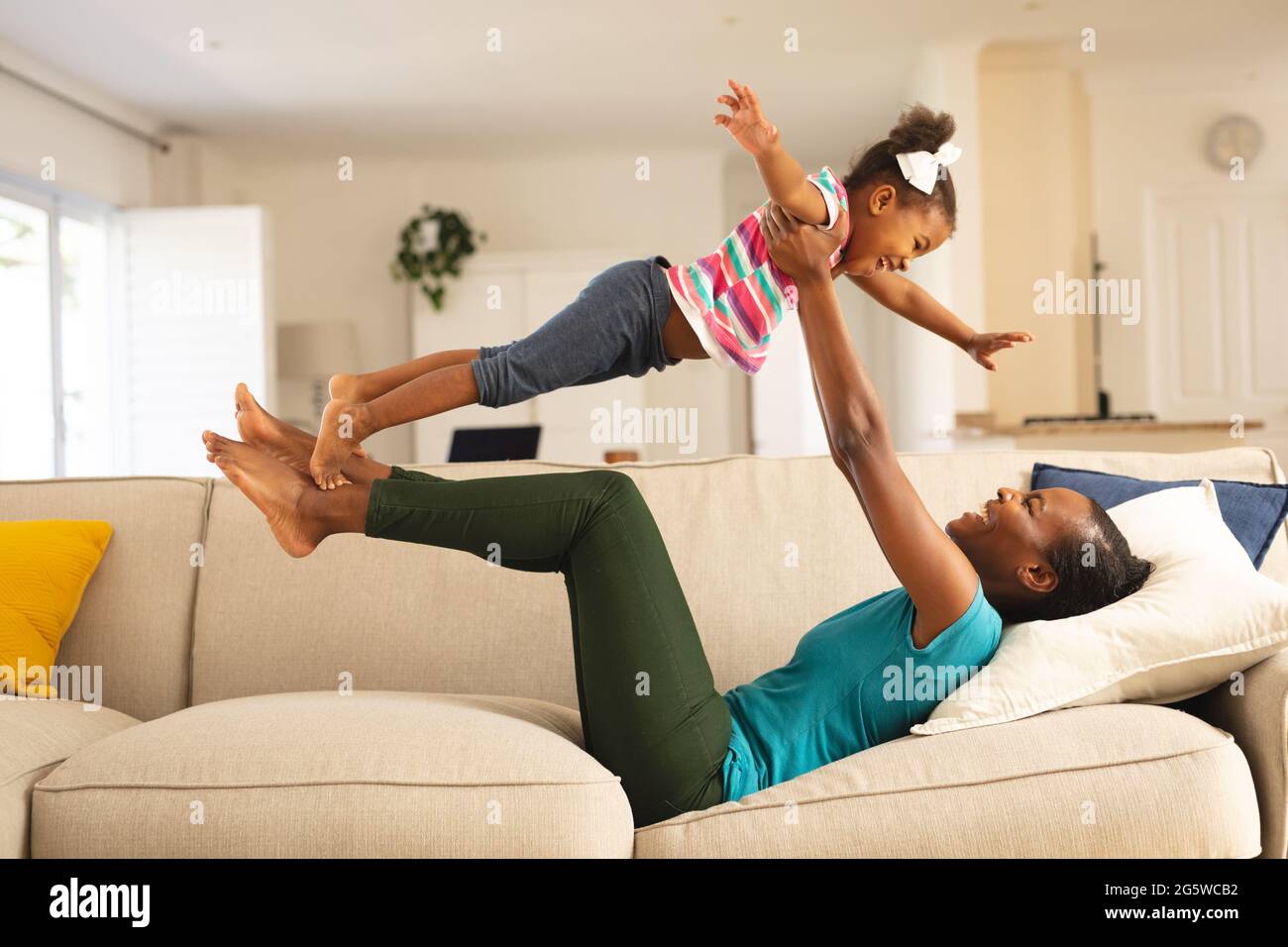 Glückliche afroamerikanische Mutter, die auf der Couch liegt und Spaß hat, lächelnde Tochter über ihr zu balancieren Stockfoto