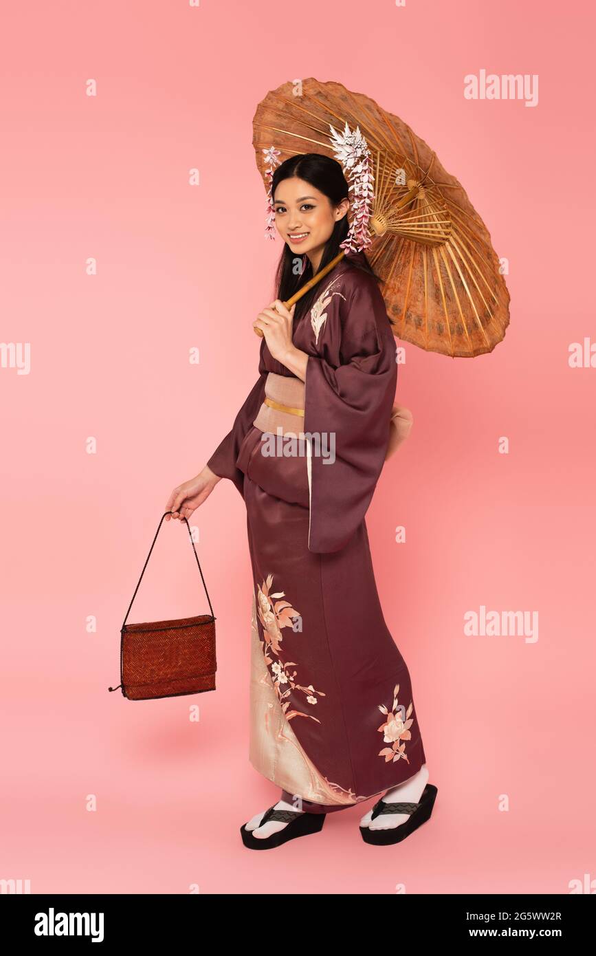 Lächelnde asiatische Frau mit Regenschirm und Handtasche auf rosa Hintergrund Stockfoto